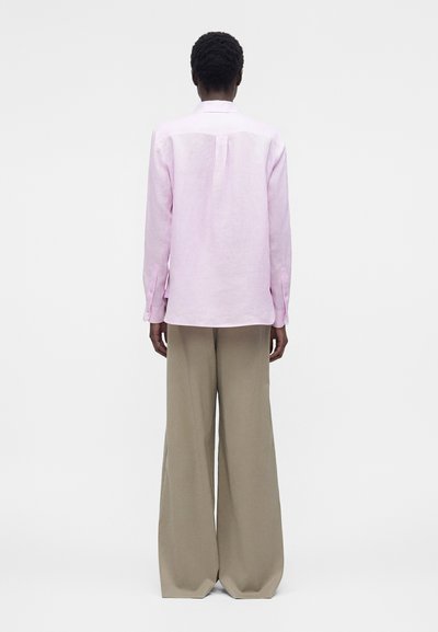 Άτομο που φαίνεται από πίσω, φοράει ανοιχτό ροζ πουκάμισο με μακριά μανίκια και φαρδιά μπεζ παντελόνια, στέκεται μπροστά σε απλό λευκό φόντο.