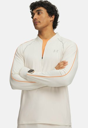 Man met gekruiste armen, die een wit shirt met lange mouwen en een kwartritssluiting draagt, met oranje biezen en een sporthorloge om zijn linker pols.