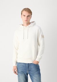 Biela mikina s kapucňou, predným vreckom a šnúrkou na stiahnutie. Vyrobená z mäkkého materiálu, s logom na ľavom rukáve. Kombinovaná s modrými džínsy.