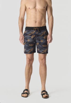 Hombre sin camisa con pantalones cortos de baño camuflaje negro y gris con patrones de tiburón naranja y sandalias negras, de pie frente a un fondo liso.