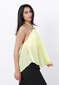 Mujer con pelo largo y oscuro que lleva un top halter amarillo claro y pantalones negros, de pie de lado frente a un fondo blanco.