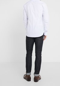 Vit långärmad skjorta med ett subtilt prickmönster, bärs med mörka jeans och bruna skor, vilket visar en enkel och figurnära design.