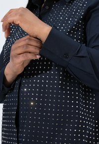 Navy shirt featuring shiny metallic studs in a pattern on the front, with a button-up collar and cuffs. The fabric appears smooth.