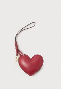 Κόκκινο δερμάτινο μπρελόκ σε σχήμα καρδιάς με λουράκι για τον καρπό και μικρό χρυσό γούρι σε ανοιχτό φόντο.