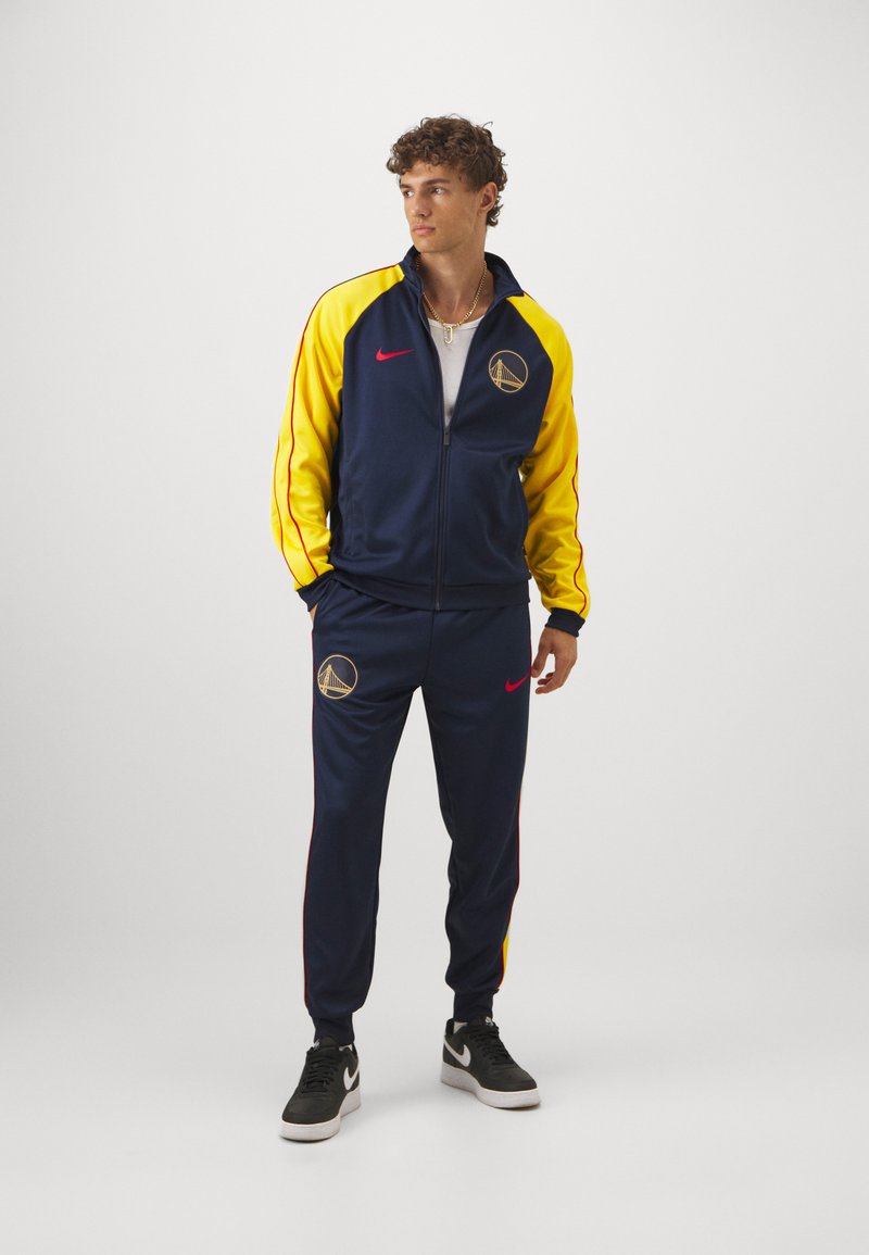 Tuta Nike navy e gialla composto da giacca con zip e pantaloni abbinati con logo, dettagli rossi e polsini a coste. Sneakers nere completano il look.