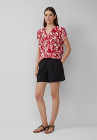 Blouse rouge et blanche à manches courtes avec un motif de feuilles, associée à un short noir et des sandales plates noires. Le tissu semble léger.