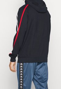 Schwarzer Hoodie mit roten und weißen Seitenstreifen, ausgestattet mit einer Kordelzugkapuze. Kombiniert mit blauen Satin-Trainingshosen, die Logodetails zeigen.