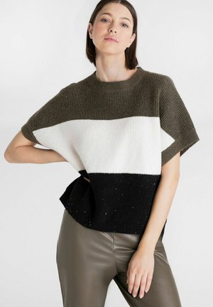 Vrouw draagt een driekleurige gebreide top met olijfgroene, witte en zwarte horizontale strepen en olijfgroene nepleren broek tegen een effen achtergrond.