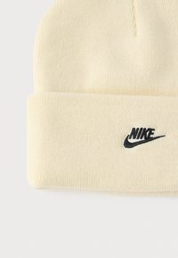Căciulă crem din material tricotat cu nervuri. Prezintă un logo Nike negru pe față. Margine pliabilă pentru ajustare și căldură.