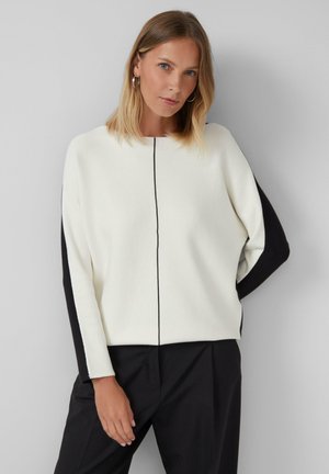 Vrouw met blond haar draagt een witte trui met zwarte mouwstrepen en een dunne zwarte middenlijn, gecombineerd met een zwarte broek, staand tegen een grijze muur.