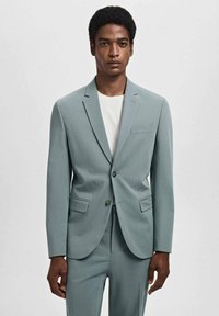 Completo turchese chiaro con giacca strutturata e pantaloni coordinati. La giacca presenta una chiusura a un bottone, rever a punta e tasche frontali.