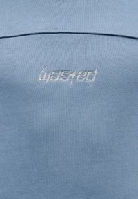 Φως μπλε ύφασμα με λευκό κεντημένο κείμενο «wasted» σε στυλιζαρισμένη γραμματοσειρά, κεντραρισμένο κάτω από μια οριζόντια ραφή.