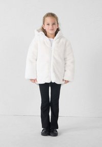 Chaqueta de piel sintética blanca con capucha, cierre de cremallera y mangas anchas. Combinada con pantalones y zapatos negros, de pie frente a un fondo neutro.