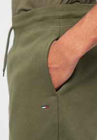 Ελιά πράσινα παντελόνια joggers φτιαγμένα από μαλακό βαμβάκι, με ελαστική μέση και πλάγιες τσέπες. Μικρή κομμένη λεπτομέρεια λογοτύπου σε κόκκινο, άσπρο και μπλε.