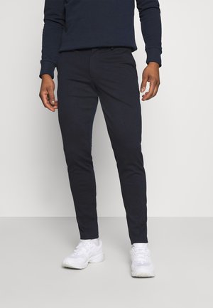 Mand iført tætsiddende sorte bukser, navy bluse med lange ærmer og hvide sneakers, stående på en lys grå gulv med en ensfarvet hvid baggrund.