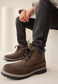 Braune Wildleder-Stiefeletten mit Schnürung, Metallösen und einem schwarzen Lederkragen. Profilierte Sohle mit tiefem Profil.