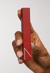 Rød rektangulær leppeprodukt holdt i en hånd, med en matt finish og minimalistisk hvitt logo. Jevn tekstur uten synlige mønstre.