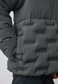 Close-up of dark gray padded jacket with subtle "BY TED BAKER" text pattern and a person’s arm bent at the side.