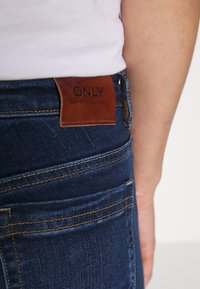 Primer plano de una persona que lleva jeans de mezclilla azul oscuro con un parche de cuero marrón etiquetado "ONLY" en la cintura y una camiseta blanca metida por dentro.