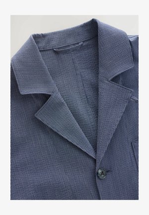 Niebiesko-biała marynarka w paski z klapami, zapinana na jeden guzik, wykonana z fakturowanej tkaniny. Posiada kieszeń piersiową oraz klapę.