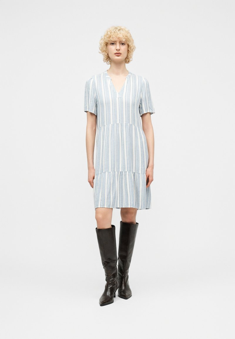 Γυναίκα με ξανθά σγουρά μαλλιά, ντυμένη με γαλάζιο και λευκό ριγέ φόρεμα μέχρι τα γόνατα και μαύρες δερμάτινες μπότες μέχρι το γόνατο, στέκεται μπροστά σε λευκό φόντο.