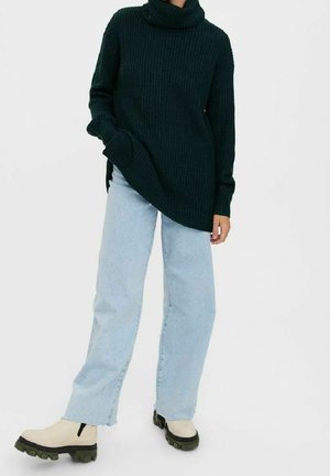 Personne portant un pull à col roulé côtelé vert foncé, un jean large bleu clair et des bottes beiges à semelle épaisse, debout devant un fond blanc.