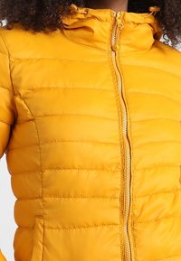 Żółta ocieplana kurtka z zamkiem z przodu, kapturem i bocznymi kieszeniami. Posiada pikowaną teksturę i gładki materiał, idealna na ciepło i komfort.