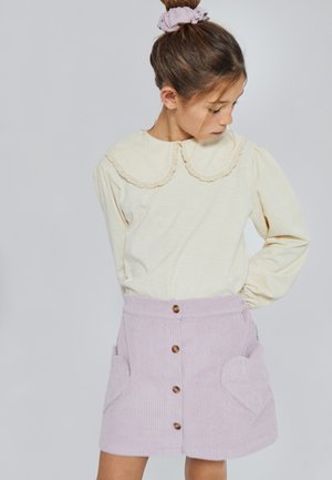 Blusa amarillo claro con mangas abullonadas y cuello con borde ondulado, combinada con una falda de pana lila con bolsillos en forma de corazón y botones.