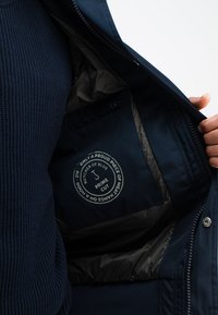 Marineblaues Jackett mit wattiertem Innenfutter und Logo-Aufnäher. Verfügt über einen Reißverschluss, eine Innentasche und eine gerippte Textur auf der Außenseite.