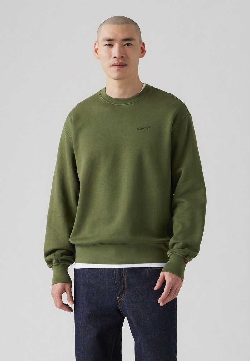 Olijfgroen sweatshirt van zachte stof, met een ronde halslijn, geribde mouwen, en een subtiele logo op de borst. Gekleed met donkere jeans.