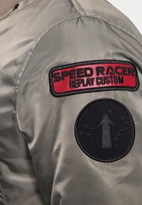Khaki bomber bunda s červenou a černou vyšívanou nášivkou s nápisem "SPEED RACER REPLAY CUSTOM" a kulatou černou nášivkou se šipkou.
