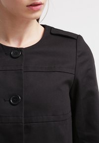 Nainen, jolla on päällä musta takki, jossa on suuret mustat napit ja hartiateipit, kuvattuna kaulasta vyötärön puoliväliin.