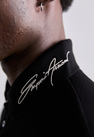 Svart polo t-shirt med en strukturerad krage som har vit broderad signaturdetalj. Närbild som framhäver textur och hantverk.