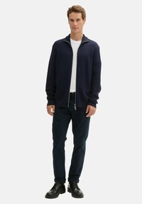 Granatowy dzianinowy kardigan z wysokim kołnierzem i srebrnym zamkiem, noszony na białej koszulce. W połączeniu z ciemnymi spodniami i czarnymi sznurowanymi butami.