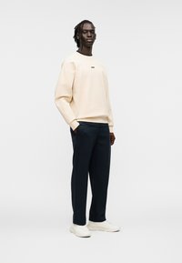 Sweat-shirt beige clair avec logo noir, associé à un pantalon bleu marine foncé et des baskets blanches. Tissu lisse avec une coupe décontractée et moderne.