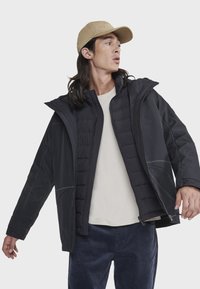 Veste noire avec une doublure rembourrée, dotée d'une capuche, de fermetures éclair et de poches latérales. Associée à un t-shirt de couleur claire et un pantalon bleu marine.