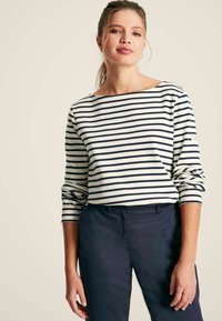 Gestreiftes langärmeliges Oberteil in Weiß und Marineblau mit weitem U-Boot-Ausschnitt. Hergestellt aus einem weichen Stoff mit hochgerollten Ärmeln. Kombiniert mit dunklen Hosen.