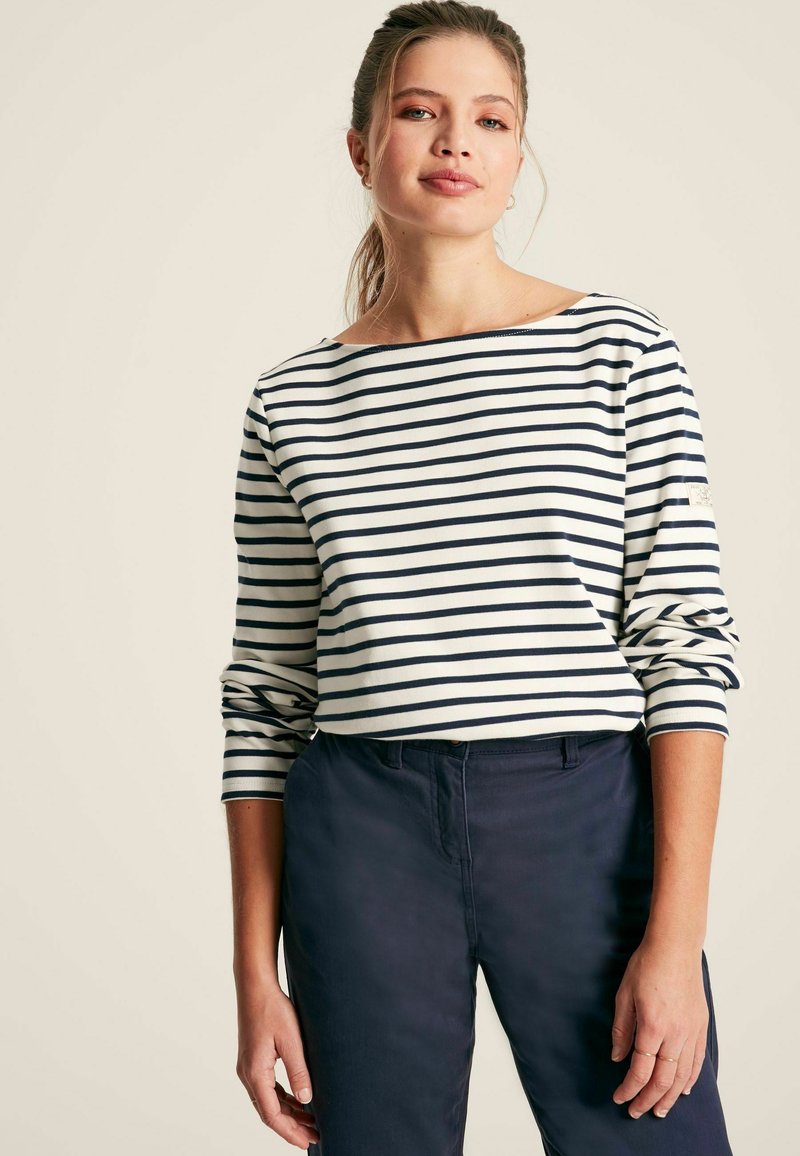 Gestreiftes langärmeliges Oberteil in Weiß und Marineblau mit weitem U-Boot-Ausschnitt. Hergestellt aus einem weichen Stoff mit hochgerollten Ärmeln. Kombiniert mit dunklen Hosen.