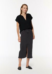 Kobieta stoi ubrana w czarny top z krótkim rękawem, ciemnoszary midi spódnicę z przednim rozcięciem oraz czarne loafersy na wsuwkę z detalem w postaci łańcucha.