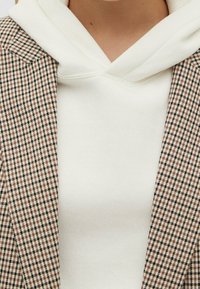 Blazer à motif pied-de-poule en marron et noir associé à un sweat à capuche blanc. Mise en avant du tissu texturé et du look superposé des vêtements.