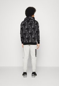 Sweat à capuche noir avec un motif texturé et un cordon de serrage, associé à un pantalon de survêtement gris avec un logo vertical, et des baskets noires.