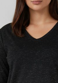 Frau mit glattem braunem Haar, die einen schwarz gesprenkelten V-Ausschnitt-Pullover trägt, der den Hals und den oberen Brustbereich zeigt.