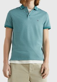 Muž v tyrkysovém polo tričku s krátkým rukávem přes bílé tričko a béžových kalhotách, stojící proti jednobarevnému pozadí.