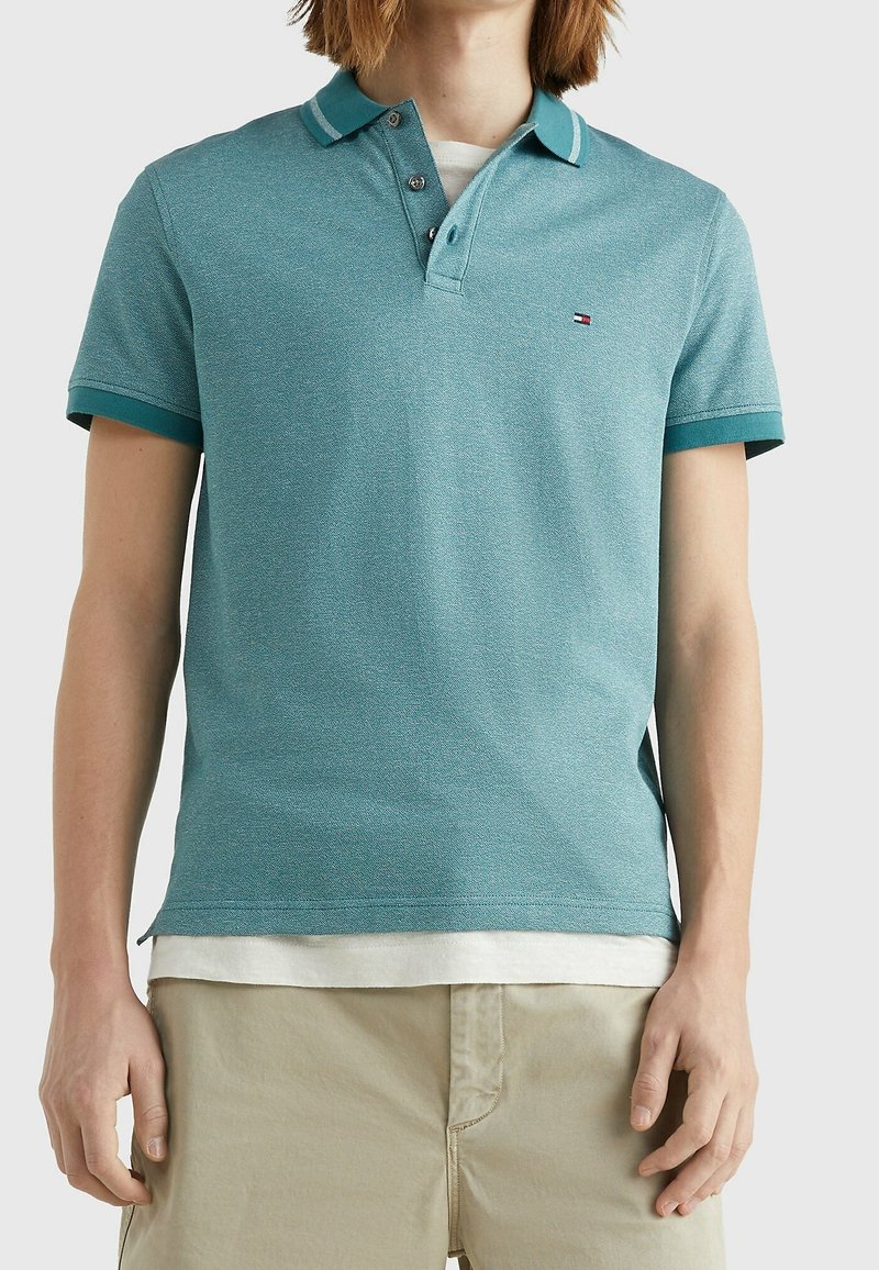 Muž v tyrkysovém polo tričku s krátkým rukávem přes bílé tričko a béžových kalhotách, stojící proti jednobarevnému pozadí.