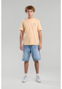 Light peach cotton t-shirt with a round neck and logo detail. Worn with light blue denim shorts, white sneakers, against a gray background.
