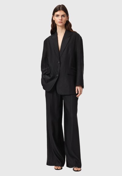 Femme debout portant un blazer noir oversize et un pantalon noir à jambes larges avec des sandales noires à brides sur un fond clair uni.