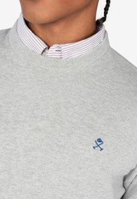 Suéter gris con cuello redondo y puños acanalados, combinado con una camisa de cuello blanca y roja a rayas. Acento de logotipo azul bordado.