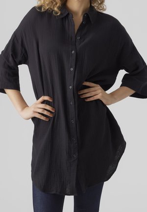 Kobieta w luźnej, teksturowanej, czarnej koszuli na guziki z rękawami 3/4, ręce na biodrach, na jednolitym jasnym tle.