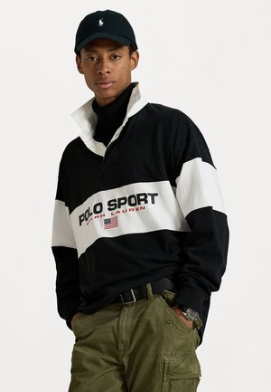 Polo noir et blanc à manches longues avec logo "POLO SPORT" et détail du drapeau américain. Porté par-dessus un col roulé noir et associé à un pantalon cargo olive.