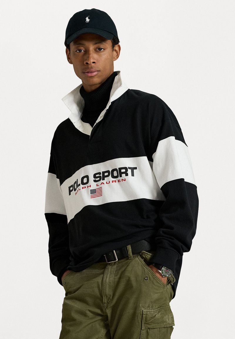 Zwart-witte langemouwen polo met het "POLO SPORT" logo en een detail van de Amerikaanse vlag. Gedragen over een zwarte coltrui en gecombineerd met olijfgroene cargobroek.
